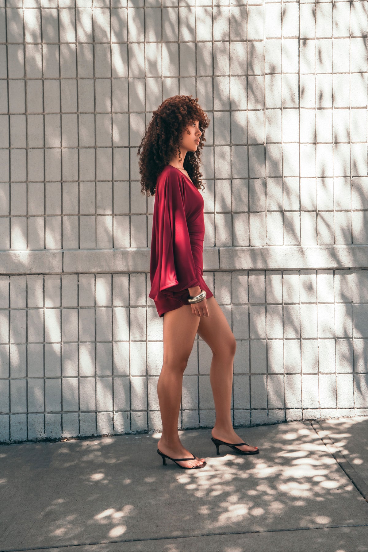 A woman with curly brown hair stands in profile against a white tiled wall with dappled sunlight and shadows. She is wearing a dark red long-sleeve top and matching shorts, paired with black heeled sandals.